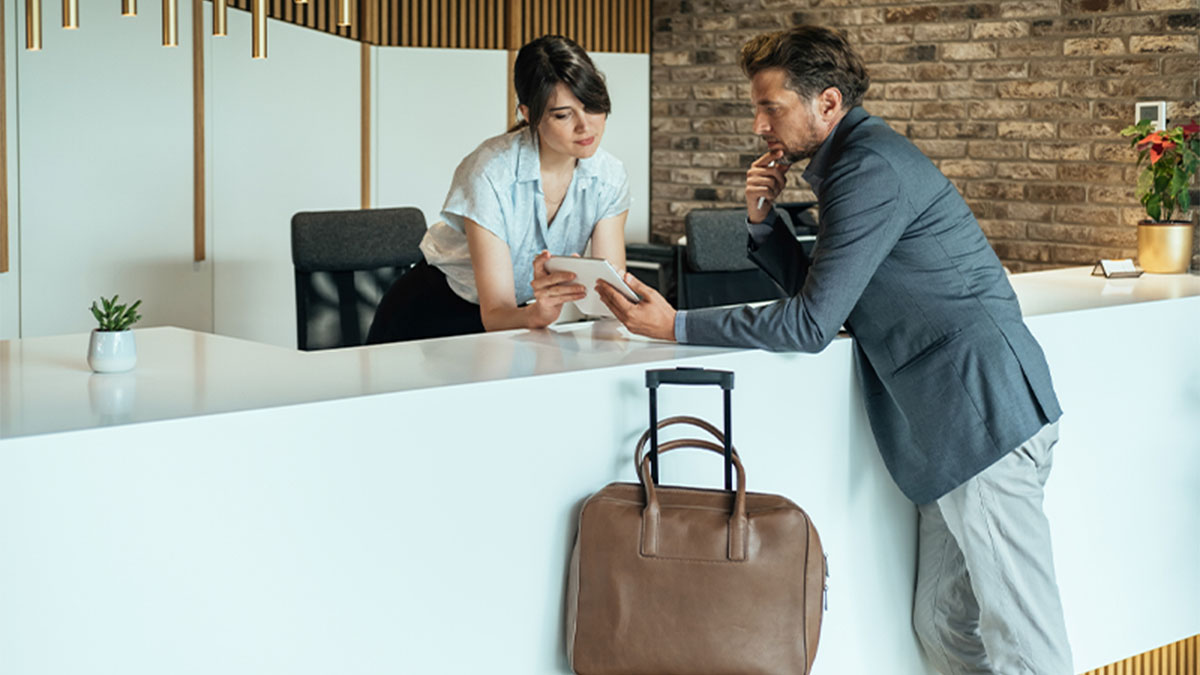 Cliente em lobby de hotel com mala de viagem a tirar dúvidas com rececionista